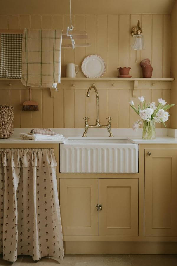 Cream fluted farmhouse sink from @nicolahuthwaite. Vintage brass faucet with buttercream yellow cabinets and spice print curtains.