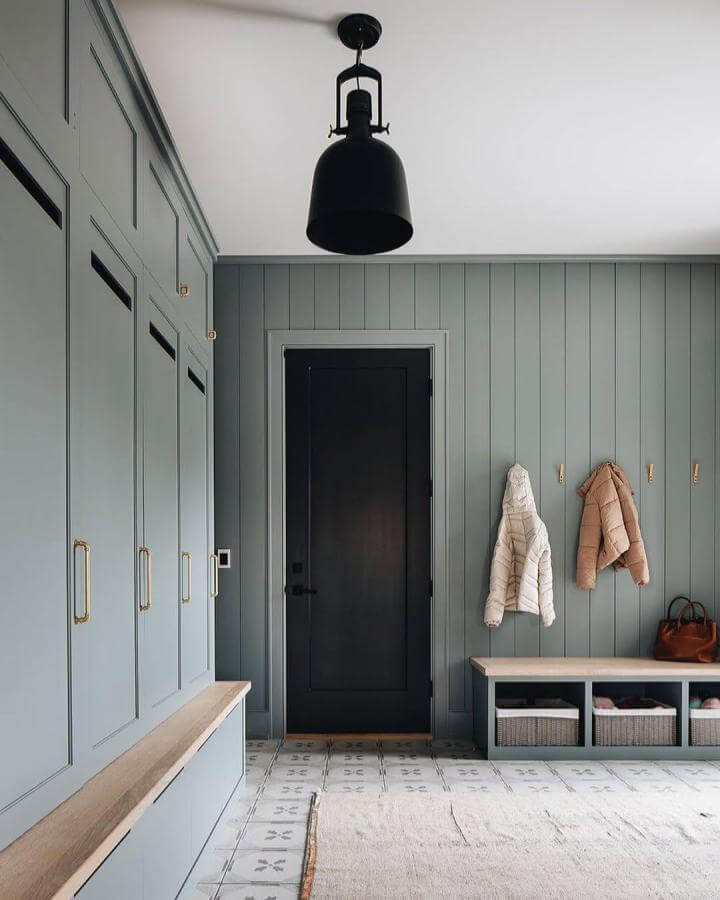 Sage green paneled mudroom with brass hardware and patterned floor tiles from @stofferphotographyinteriors. Light wood bench and hooks for coats.