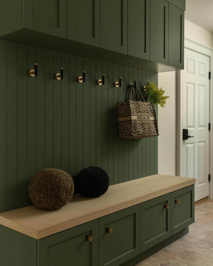 Sage green paneled mudroom bench and cabinets from @raquellangworthy.photo with gold hooks and natural wood countertop.