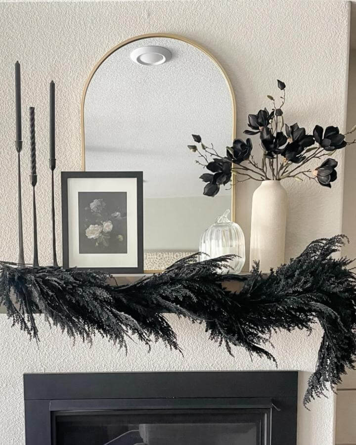 Black garland mantel display from @myneutralhomeca. Moody foliage drapes across white fireplace with candles and neutral decor.