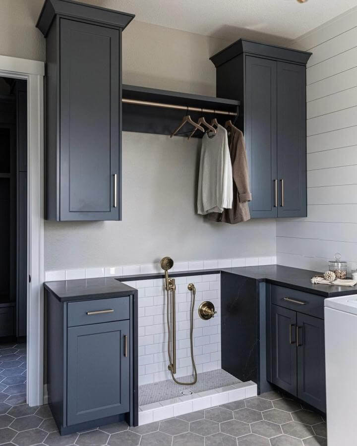 Dark gray cabinetry and white tiled dog washing area with brass fixtures in a stylish mudroom from @juniperandjadeinteriors offering a functional pet wash spot.