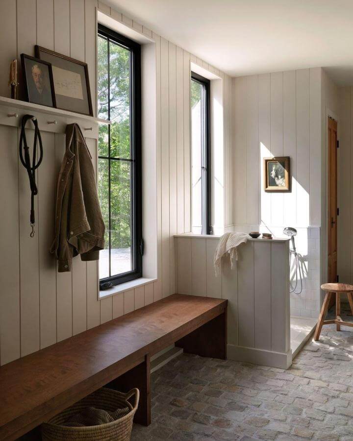 Warm wood bench and rustic brick floored mudroom with black window frames and white paneled walls by @sylvieli designed for pet cleanup.