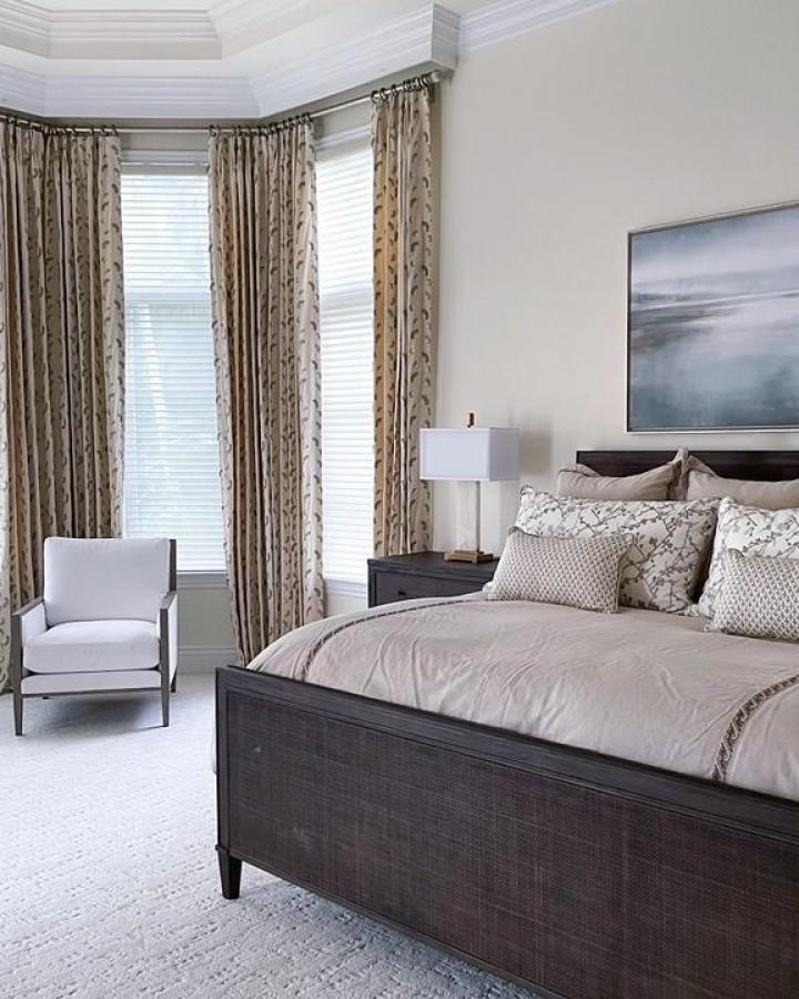 Neutral bedroom with corner windows and patterned curtains from @sandraasdourianinteriors . Dark wood furniture complements beige color scheme.